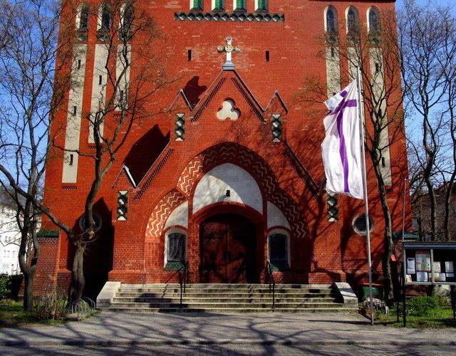 Rote Backsteinkirchenfassade mit weiß-lila Fahne.